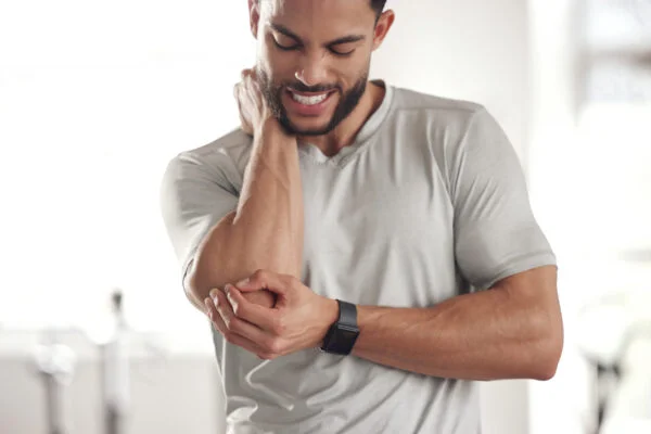 Athletic man in a t-shirt wincing while gripping his aching elbow joint, representing targeted SoftWave therapy treatment for chronic elbow pain.