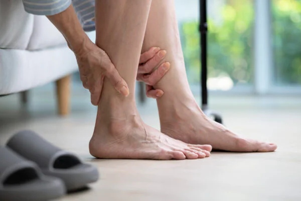 Close-up of lower legs and bare feet on the floor with a person holding their ankle in pain, representing common foot and ankle conditions manageable with non-invasive SoftWave therapy.